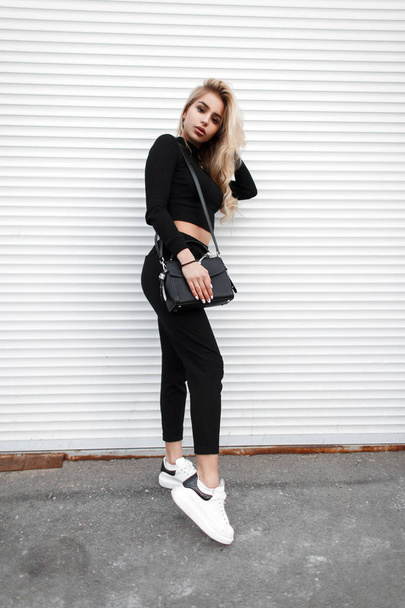 Beautiful young stylish blond woman in black fashion clothes with white shoes posing on the street - Photo, Image