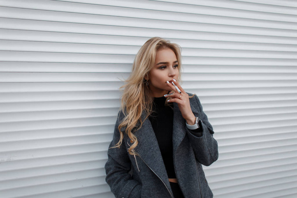 Élégante belle jeune femme dans une veste de mode grise avec une cigarette près d'un mur en métal blanc dans la rue
 - Photo, image