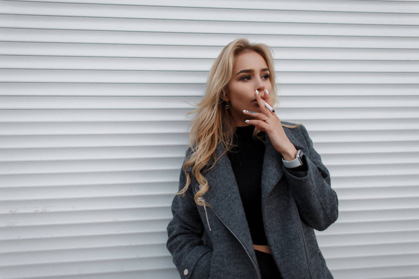 Beautiful young model woman with a cigarette in a gray fashionable jacket near a white metal wall in the street - Foto, Imagem