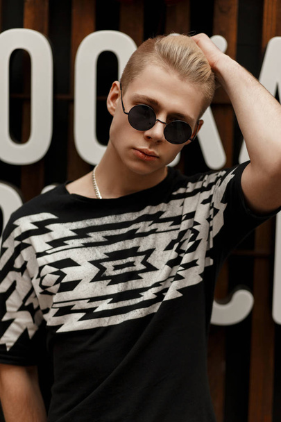 Élégant jeune homme modèle avec une coiffure en lunettes de soleil vintage rondes dans un T-shirt de mode noire avec impression se tient près d'un mur en bois avec des lettres
 - Photo, image
