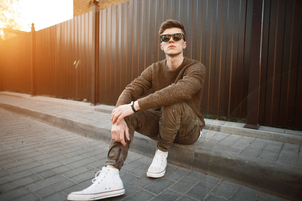 Handsome fashion hipster man in stylish sunglasses in a fashionable pullover and military stylish pants with white shoes sits on the asphalt on the street at sunset - Photo, Image
