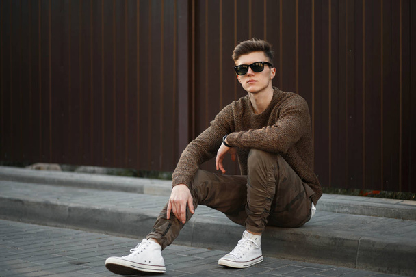 Стильный модный молодой человек в модной одежде сидит на улице возле металлической стены
 - Фото, изображение
