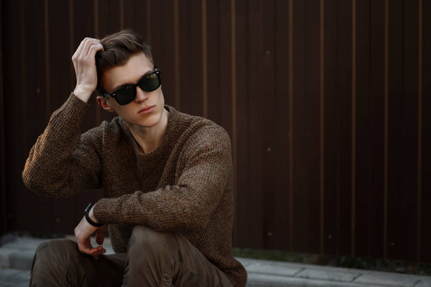 stylischer junger Mann mit Sonnenbrille und Frisur sitzt an einer Metallwand auf der Straße - Foto, Bild