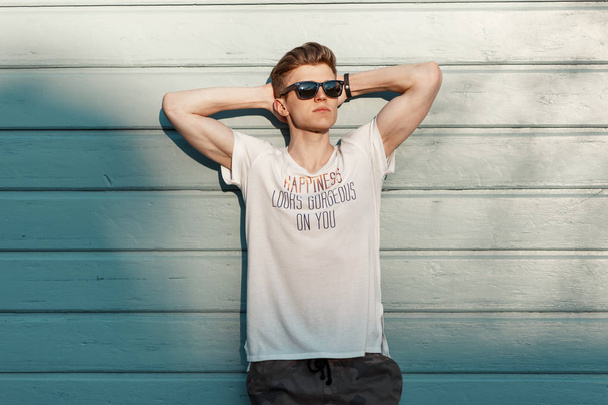 Guapo joven hipster hombre con gafas de sol en ropa de moda descansando cerca de una pared de playa de madera azul en un día soleado
 - Foto, imagen