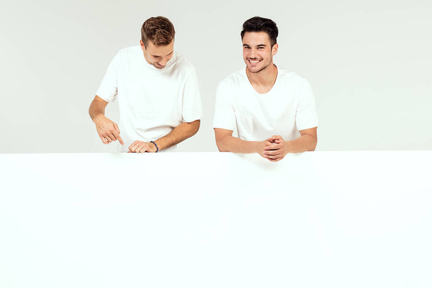 Two young handsome guys holding blank white banner, signboard, isolated on white studio background. - Zdjęcie, obraz