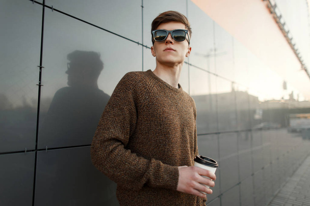 Young handsome man with coffee in sunglasses and fashionable clothes stands near the wall at sunset - 写真・画像