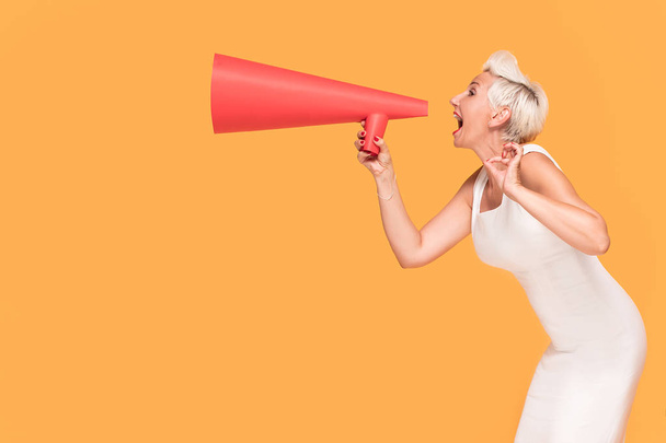 Middle age elegant woman shouting loud by red megaphone, smiling, looking at camera. Lady expressing positive concept on orange studio background. - Photo, Image