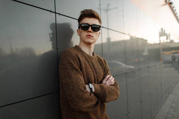 Handsome young hipster man in sunglasses in trendy stylish clothes stands near a modern wall - Photo, Image