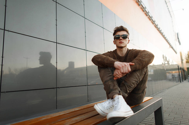 Stylish handsome young man with sunglasses in a knitted pullover and trendy white sneakers with fashion military pants sitting on a bench in the city - Φωτογραφία, εικόνα