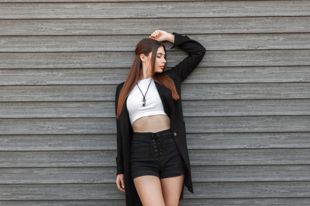 Menina modelo jovem bonita com um penteado em um casaco na moda preto com shorts posando perto de uma parede de madeira. Rua moda moda moda
 - Foto, Imagem