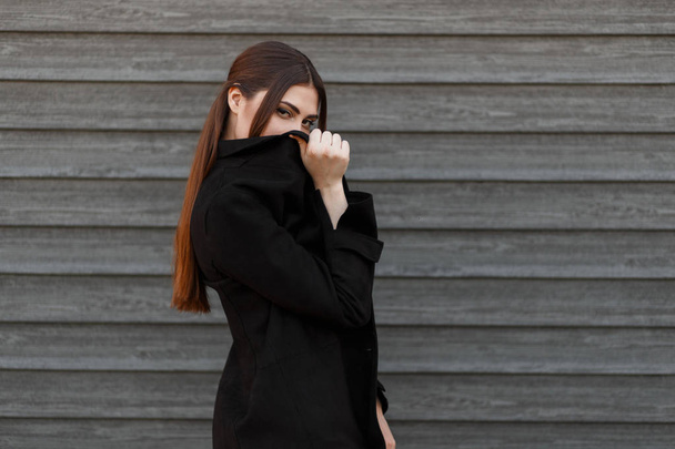 Μοντέλο γυναίκα σε ένα μαύρο παλτό καλύπτει το πρόσωπό της κοντά σε ένα ξύλινο τοίχο - Φωτογραφία, εικόνα