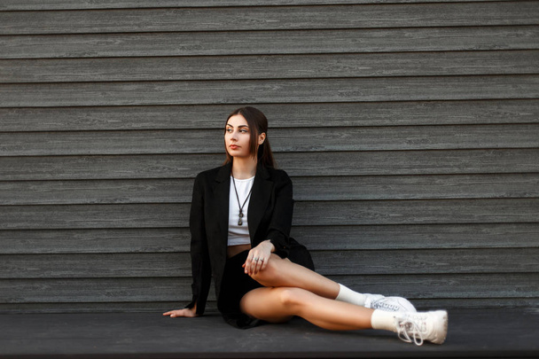 Молодая красивая стильная женщина в черном пальто с белыми модными кроссовками отдыхает у деревянной винтажной стены
 - Фото, изображение