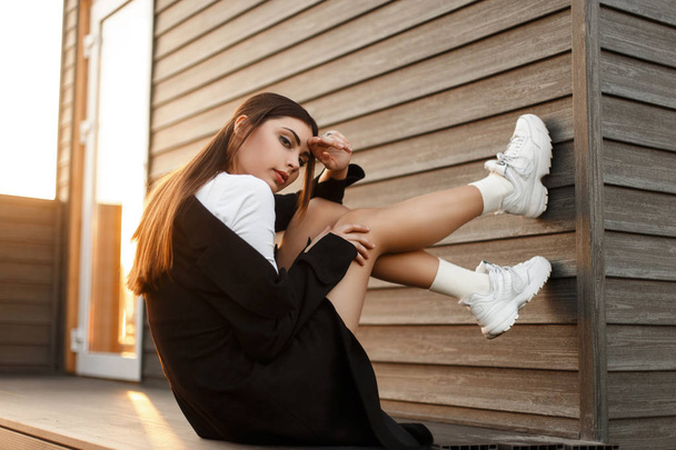 Όμορφο νεαρό μοντέλο μόδας γυναίκα στο μαύρο κομψό παλτό με πάνινα παπούτσια κάθονται κοντά σε ένα ξύλινο τοίχο στο ηλιοβασίλεμα - Φωτογραφία, εικόνα