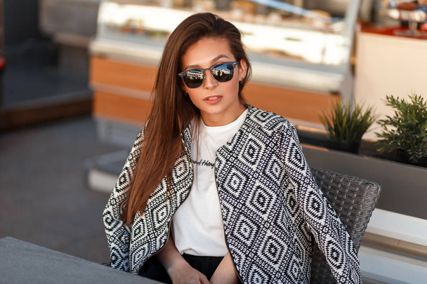 Hezká mladá žena stylový model v módní bunda s sluneční brýle sedí v restauraci na ulici - Fotografie, Obrázek