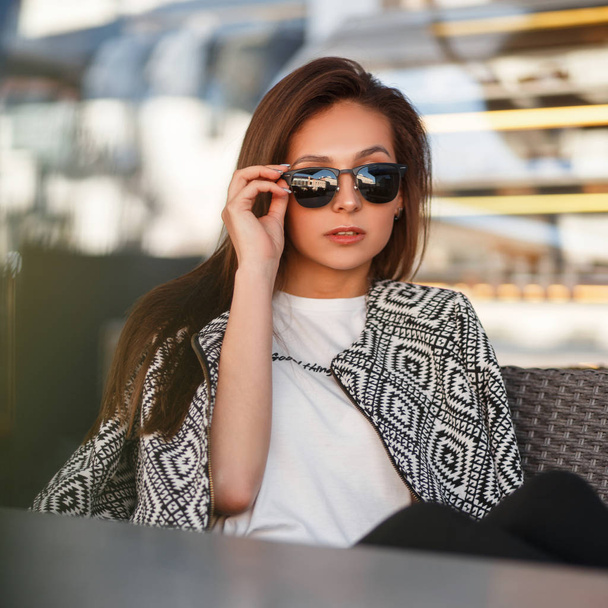 Beautiful young model woman with sunglasses in a white T-shirt with a fashion bomber jacket sits to a cafe on the street - Photo, Image
