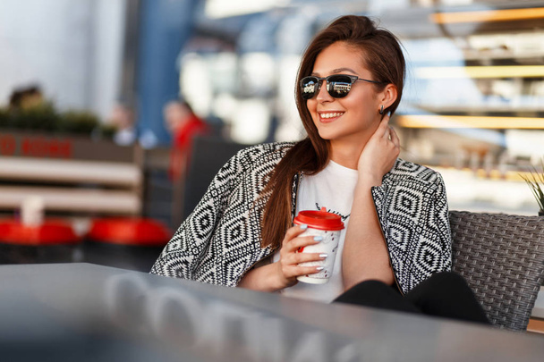 Красива модна молода жінка з кавою в сонцезахисних окулярах зі стильним модним одягом відпочиває в ресторані на вулиці
 - Фото, зображення