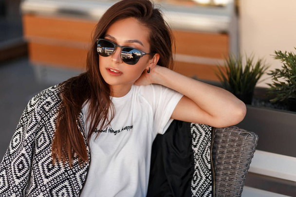 Muodikas kaunis nuori nainen muoti aurinkolasit tyylikäs pommikone takki ja valkoinen t-paita istuu kahvilassa kadulla
 - Valokuva, kuva
