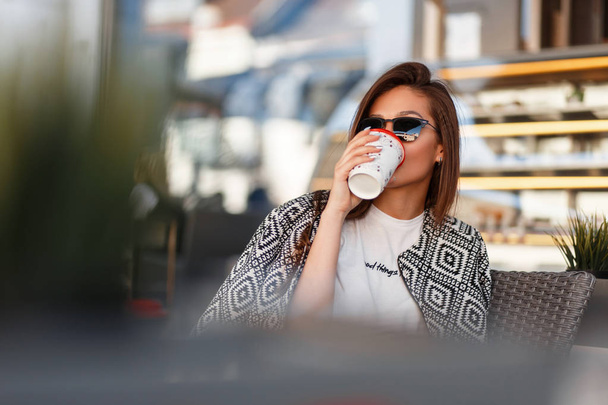 Модна красива молода жінка в стильному одязі п'є каву в кафе на вулиці
 - Фото, зображення
