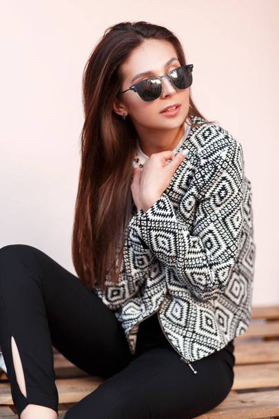 Giovane bella donna modello con occhiali da sole in una giacca elegante con modelli si siede su pallet di legno
 - Foto, immagini