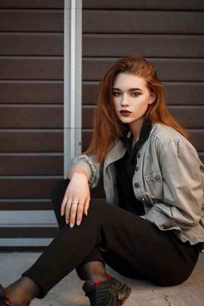 Beautiful young model woman in fashionable jacket and black T-shirt with pants sitting on the street near the gate - Fotografie, Obrázek