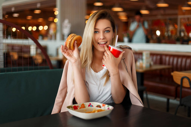 Happy funny young woman with a hamburger eating fast food in a cafe - Foto, immagini