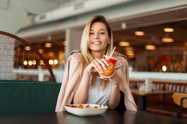 Happy young beautiful woman in pink jacket with fast food, hamburger and cola sitting in cafe - Photo, Image