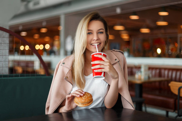 Beautiful young woman with a hamburger drinks a cola in a cafe - Photo, Image