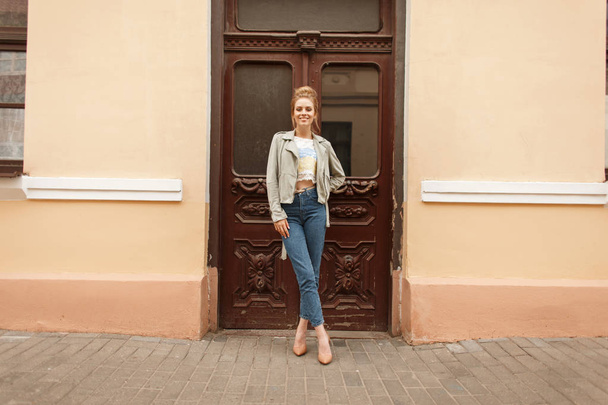 Fashionable young model girl in vintage fashion jacket with jeans posing near old wooden doors on the street - Foto, Imagen