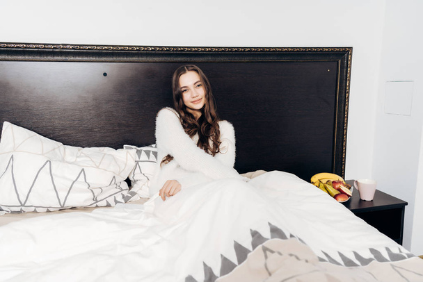 charming young girl in white pajamas lies in bed, woke up early in the morning in a good mood - Foto, Imagem