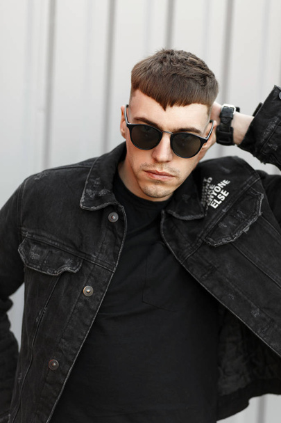 schöner junger Mann amerikanisches Model mit einer Frisur in Sonnenbrille in schwarzer modischer Jeanskleidung auf der Straße - Foto, Bild