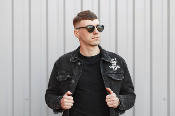 Модний молодий чоловік з сонцезахисними окулярами в чорному джинсовому одязі позує на вулиці біля сірої металевої стіни
 - Фото, зображення