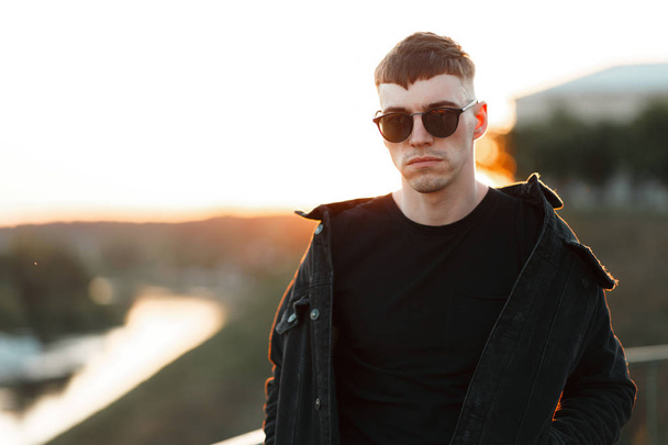 stylish young handsome man with sunglasses in black fashionable clothes at sunset - Фото, зображення