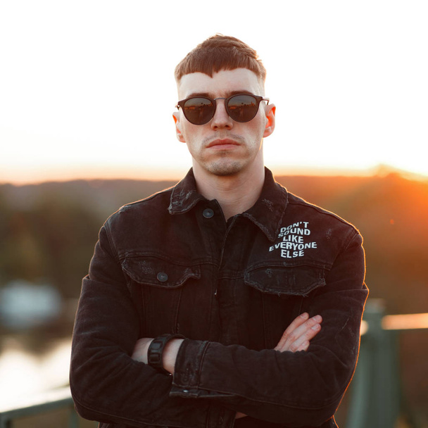 Fashionable young man with stylish sunglasses in a black denim jacket at sunset - Фото, зображення