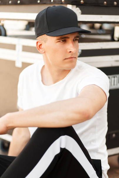 Handsome young model man with a cap in stylish black and white clothes sitting on the street - Photo, Image