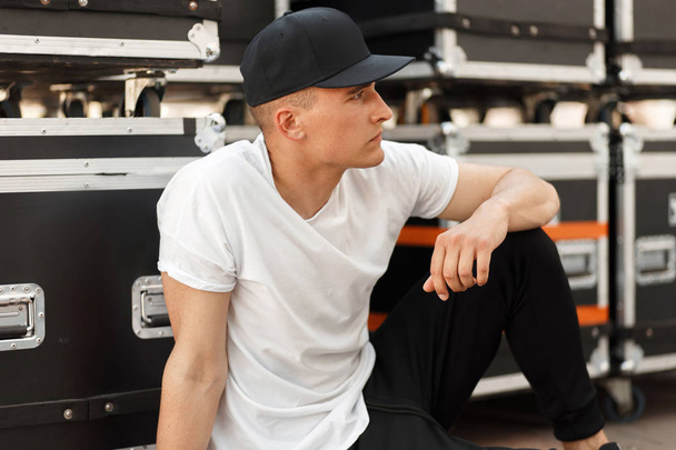 muodikas nuori mies musta lippis ja valkoinen T-paita istuu lähellä kuljetuslaatikot kadulla
 - Valokuva, kuva
