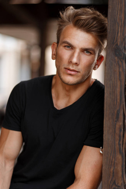 Handsome young male model with a hairstyle in a black stylish T-shirt near a wooden pillar outdoors - Foto, Bild
