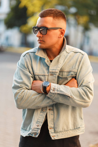 Hipster hombre con un peinado elegante en gafas de sol y ropa de mezclilla de moda en la ciudad
 - Foto, imagen