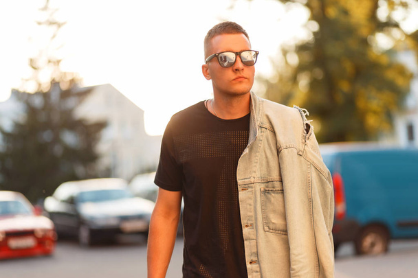 Fashionable man with stylish sunglasses in a black T-shirt and jeans jacket at sunset - 写真・画像