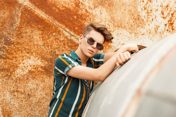 Fashionable handsome guy with a stylish hairstyle with sunglasses in a summer color shirt near the metal rusty technique - 写真・画像