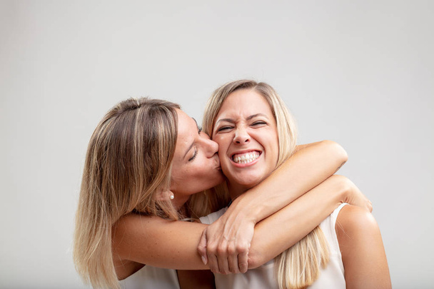 Affectionate young blond woman hugging her friend or partner and kissing her on the cheek in a close up view on white - Photo, Image