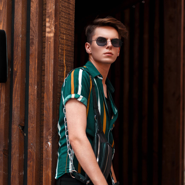 Stylish handsome young man with trendy sunglasses in fashion beach clothes near a wooden wall - 写真・画像