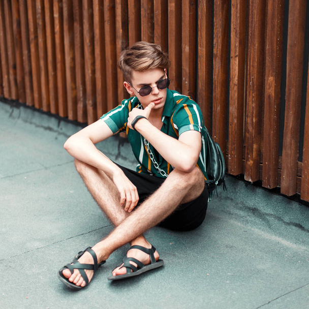 handsome stylish man with stylish sunglasses in a summer shirt with shorts and a purse sitting near the wooden wall - Photo, Image