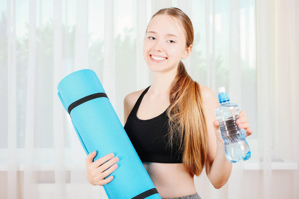 Meisje met blauwe gymnastiekmat en fles water voor raam. Concept sport. - Foto, afbeelding