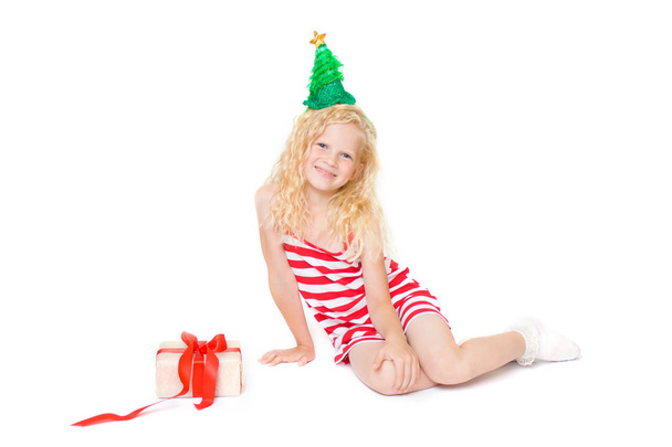 holidays, winter and celebration concept -Christmas and new year card with cute little blonde girl in red and white carnival costume dress and green fir tree hat with gift box isolated on white - Foto, immagini