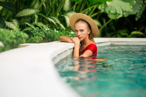 Retrato de moda de uma bela jovem mulher em maiô vermelho sexy e chapéu de palha vintage posando na água da piscina azul. Um dia de sol quente. Férias na ilha tropical. Menina de viagem de verão, estilo de vida hipster ativo
. - Foto, Imagem