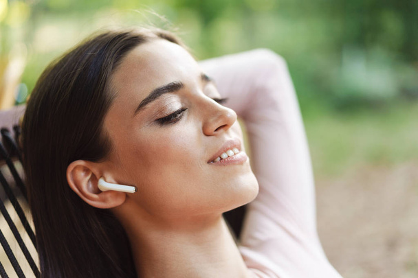 Close up of a smiling young brunette girl in wireless earphones relaxing on a chair outdoors - Foto, imagen