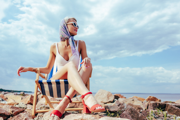 岩の多い海岸にビーチの椅子に座っているレトロな水着でエレガントな女性 - 写真・画像