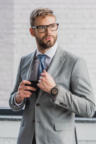 handsome young businessman in suit and eyeglasses holding smartphone and looking away - Photo, Image