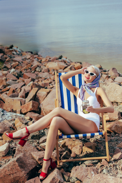 happy beautiful girl holding lemonade and resting in beach chair on rocky shore - Φωτογραφία, εικόνα