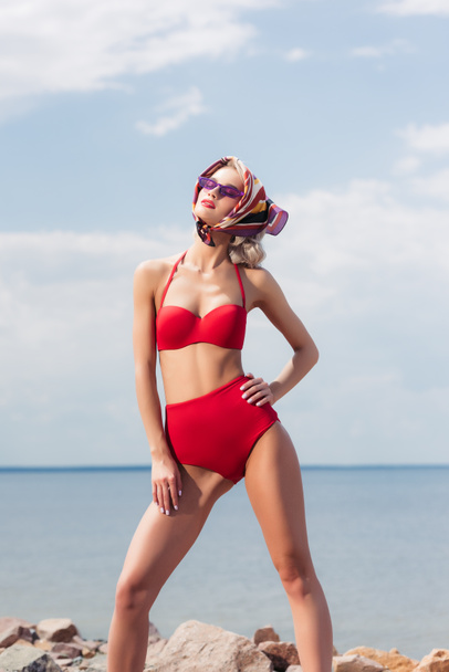 stylish elegant girl posing in red bikini, silk scarf and sunglasses on rocky beach - Photo, Image